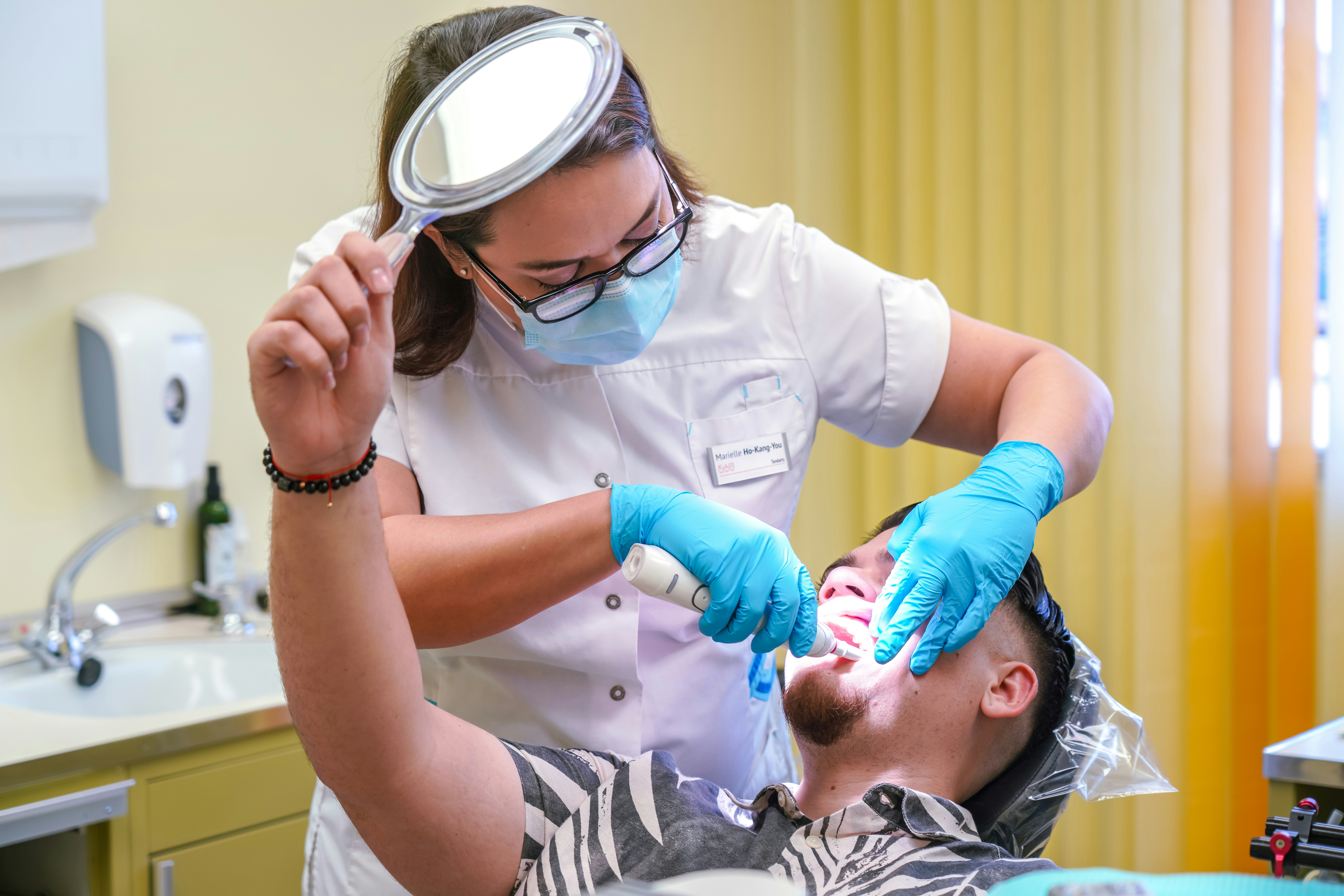 female dental assistant working on patient's teeth while he holds mirror